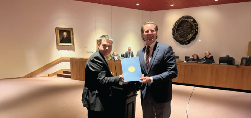 Foreground: Brendan Madden is on the left is dressed in a dark suit and is holding one side of a blue folder with an official-looking gold seal.  Deaglan McEachern is on the right, also in a suit and tie, is holding the other side of the folder. Both are smiling slightly, suggesting a ceremonial or official presentation.  Background: Several people are seated behind a long wooden desk, resembling a city council or government meeting setup.  The room has beige walls, a reddish-brown ceiling, and official decor, including a large circular seal on the wall and a portrait of a man in historical attire.  There is also a staircase and elevated platform on the left side of the room.  The setting, along with the blue folder bearing a seal, suggests that this is the presentation or receipt of a proclamation.