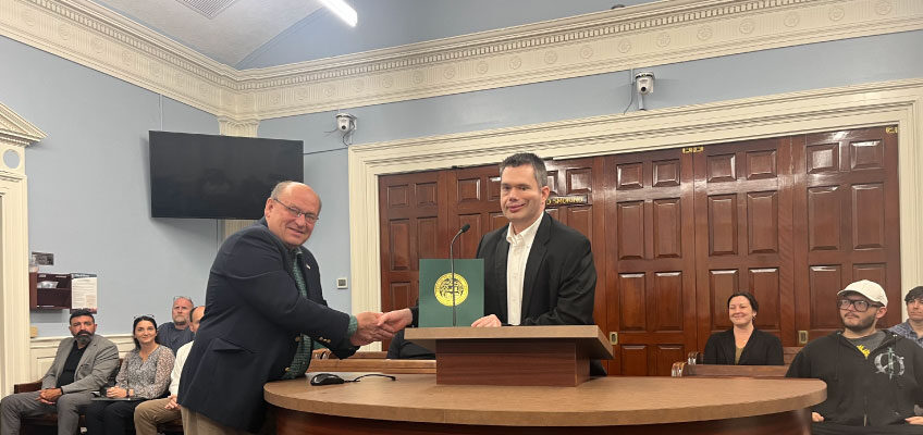 The image shows a formal setting, likely inside a courtroom or a government chamber. Two men are at the podium in the foreground. Mayor Robert Carrier is on the left, wearing glasses, a dark jacket, and a tie, is leaning forward and shaking hands with the man on the right. He is smiling.  Brendan Madden is on the right, dressed in a black suit jacket and white shirt, stands behind the podium. In front of him is a folder or certificate with a seal/logo on it. He is also smiling.  Behind them, there are wooden double doors and a group of people seated, watching the exchange. Some are smiling as well.  The room has ornate molding along the ceiling and a large flat-screen TV mounted on the wall to the left.  The scene suggests a recognition, award, or formal presentation ceremony taking place in a municipal or government setting.