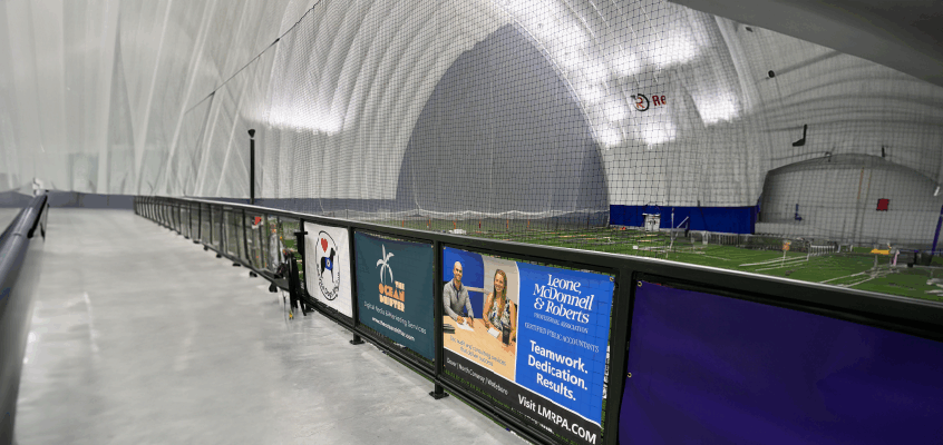 The image shows the inside of a large indoor sports facility, likely a sports dome. The structure has a high, arched, fabric-like ceiling supported by air pressure or framing. A net runs along the right side, separating a walkway from a field area. On the field side, you can see artificial turf with lines marked for sports like soccer or football. There are also cones and small barriers set up, possibly for drills or training. Along the railing of the walkway are several advertisements: The first banner shows a logo with a black dog in profile, wearing a blue service dog vest, and a red heart over it and below it says Service Dogs NH  The second banner advertises “The Ocean Drifter” with a palm tree logo and mentions digital media and marketing services.  The third banner is for Leone, McDonnell & Roberts, Certified Public Accountants, with a slogan about teamwork, dedication, and results.  The walkway itself has a polished concrete floor, black railings, and emergency equipment (fire extinguisher) attached to the railing. The environment looks clean, modern, and well-lit.