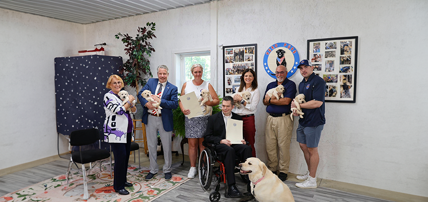 The image shows a group of seven adults gathered together indoors, holding small, five week old Yellow Labrador Retriever puppies. In the foreground,Brendan Madden is seated in a wheelchair and there is a larger, fully-grown Labrador sitting on the floor. Behind him on the left is Laura Barker, founder of Hero Pups and behind him on the right is the governor of New Hampshire, Kelly Ayotte. The room has white walls and a patterned floor. On the wall behind the group, there are two large framed photo collages and a circular logo that says "Hero Pups" with an image of a dog in the center. Two of the people in the photo are holding certificates. The scene looks celebratory and warm, at the Hero Pups facility, which provides service dogs for veterans and first responders.