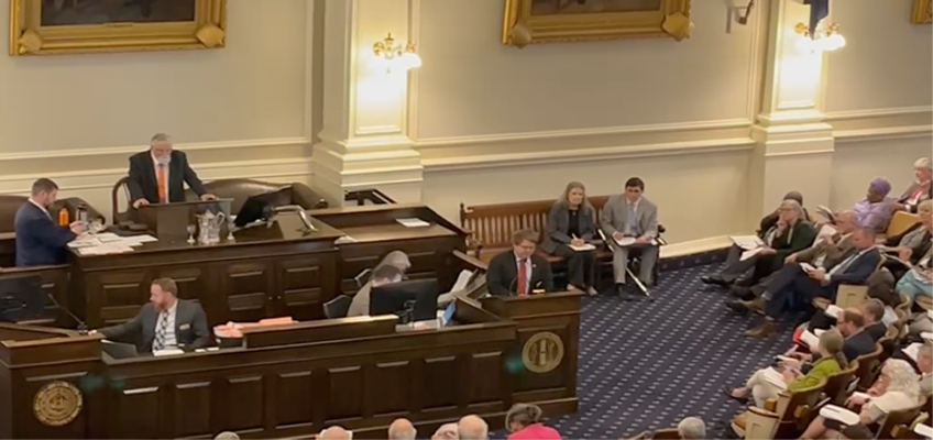 This image shows the floor of the New Hampshire House Chambers At the front, Representative Patrick Long stands at a large wooden podium or speaker’s desk, elevated above the others, overseeing the proceedings. Several other individuals are seated at desks in front of him, working with papers and computers. On the right side of the image, two people sit on a bench against the wall, appearing to listen attentively. More people are seated in rows facing the front, resembling an audience or assembly members. The room is formal, decorated with large framed paintings, tall white walls with ornate molding, and elegant light fixtures. The floor is carpeted in a dark blue pattern.