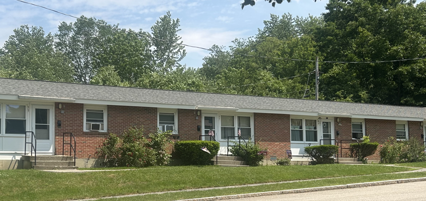 The image shows a single-story brick apartment or housing complex. The building has multiple connected units, each with its own front door and windows. Small sets of concrete steps with metal railings lead up to the entrances. Some windows have air conditioning units installed. There are bushes and small plants along the front of the building, with an American flag placed near a few of the doorways. The building has a light gray shingle roof. In the background, tall green trees are visible, suggesting the housing is in a suburban or semi-rural area. The foreground shows a grass lawn leading down to a paved road. The overall appearance is neat and well-maintained.