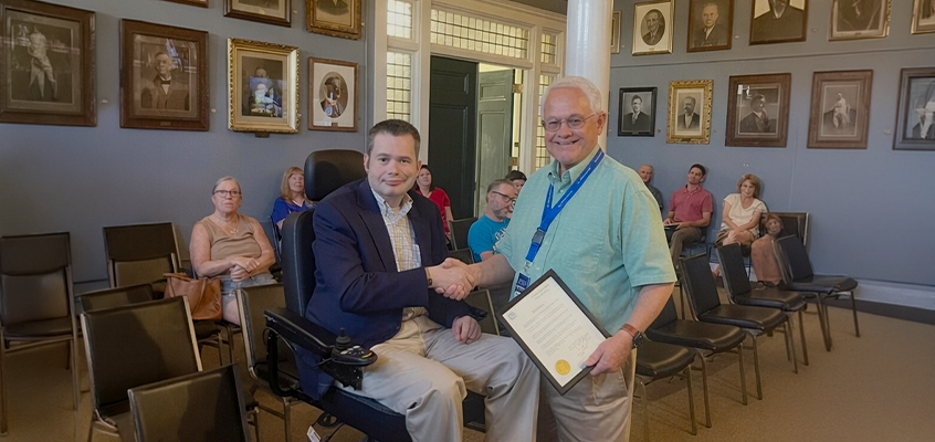 In the foreground, two men are shaking hands and smiling: • On the left is Brendan Madden, seated in a black motorized wheelchair. He has short dark hair and is wearing a navy blue blazer over a light-colored plaid button-down shirt with khaki pants. • On the right is Mayor Chuck Grassie, an older man with white hair and glasses. He is wearing a light teal/green short-sleeved collared shirt and a blue lanyard with an ID badge. In his left hand he holds a framed certificate or proclamation that features a prominent gold embossed seal (matching the City of Rochester seal from the previous image). This takes place in the Rochester City Hall Chambers, the setting is a formal indoor room with light blue walls, white columns, and numerous framed historical portraits (mostly of men in formal attire) hanging on the walls. Rows of black chairs are arranged, with several people seated in the background watching the event.