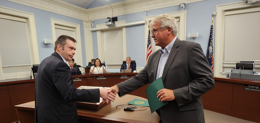 This is a photograph taken inside the Dover city council chamber .Foreground: Two men are shaking hands and smiling at each other. On the left is a younger man, Brendan Madden, with short dark hair, wearing a dark suit jacket. On the right is an older man with white/gray hair and glasses, Mayor Dennis Shanahan, wearing a gray blazer over a light blue button-down shirt. He is holding a green folder or document in his left hand. Background: A raised wooden dais/desk where several people (apparently city officials or council members) are seated. Light blue walls with white architectural molding and columns. An American flag and another flag (likely the New Hampshire state flag) standing behind the dais. A ceiling-mounted projector and other meeting room equipment are visible. Nameplates can be seen on the desks.