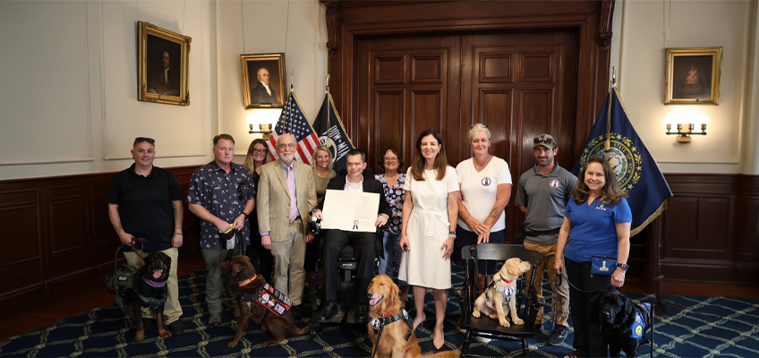 This is a formal group photograph taken indoors in an elegant, historic-looking room the executive chambers at the New Hampshire State House, Setting details: Dark wood double doors in the background. Two framed oil portraits of men in historical clothing hanging on the wall. An American flag and a dark blue New Hampshire state flag (with the state seal and “STATE OF NEW HAMPSHIRE” visible). Patterned blue carpet and classic wooden furniture (including a black Windsor-style chair). People: A mixed group of about a dozen adults of various ages is posed together. Starting from the left is Faith Harris founder of walking with honor, to her right is Sean an Operation Delta Dog client, to his right and behind him is Riley Stewart from Operation Delta Dog, to her right is Senator David Watters, dressed in a tan suit and pink tie, to his right and behind is Lynn George from Mobility Danes of New England. In the center is Brendan Madden seated in a powered wheelchair, smiling and holding up the proclamation with a blue-and-white ribbon attached. To his right and behind stands Tracy McNichol, director of operations for Mobility Danes of New England, Governor Kelly Ayotte stands to his right dressed in a white dress. To her right stands Laura Barker from Hero Pups, standing next to her is a volunteer from Hero Pups, Brady, standing to his right is Kathy Metz from Assisted Canine Training Services. Five service dogs are in the foreground, all wearing harnesses and identification vests: Nea, A black-and-tan Rottweiler, an ambassador dog for walking with honor, stands on the far left in front of Faith Harris. Luke, A chocolate Labrador stands in front of his teammate Sean. Benny, A golden Retriever from Hero Pups sitting near the center. Red, A yellow Labrador Service dog puppy in training sitting on a chair. Magnus, A black Labrador from ACTS stands in front of Kathy Metz on the far right.